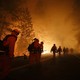 A line of firefighters walks down a road as flames loom in the background.