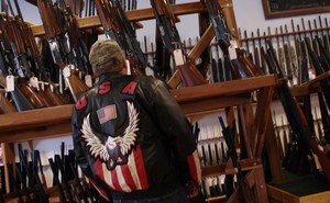 A man in a jacket featuring a bald eagle and the American flag stands in front a collection of rifles.