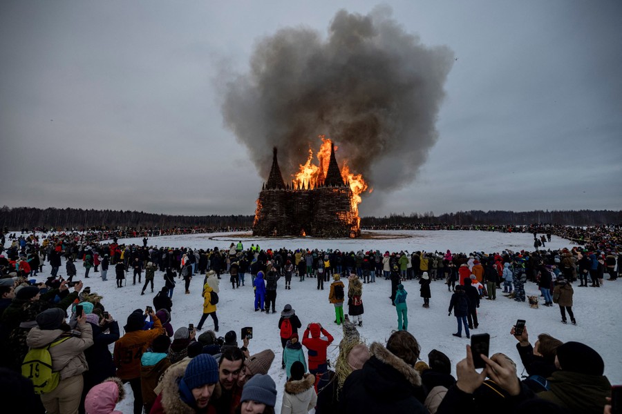 A large ring of spectators stands around a burning makeshift castle in a snow-covered field.