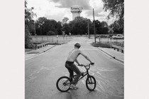 a black and white photograph of a teen boy on a bike near a water tower