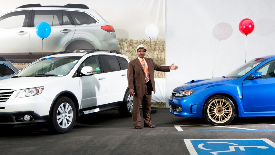 A car dealer in a brown suit holding his arm outstretched toward a blue Subaru with a red balloon tied to it