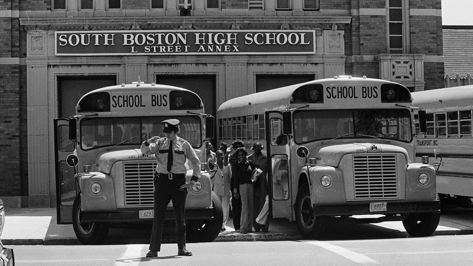 In a black-and-white photo from 1975, black students board school buses lined up in front of South Boston High School. A police office stands in front of one of the busses. 