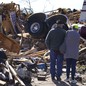 A couple stands close together, surveying extensive damage from a tornado, including a tractor unit of a semitruck atop a pile of debris.