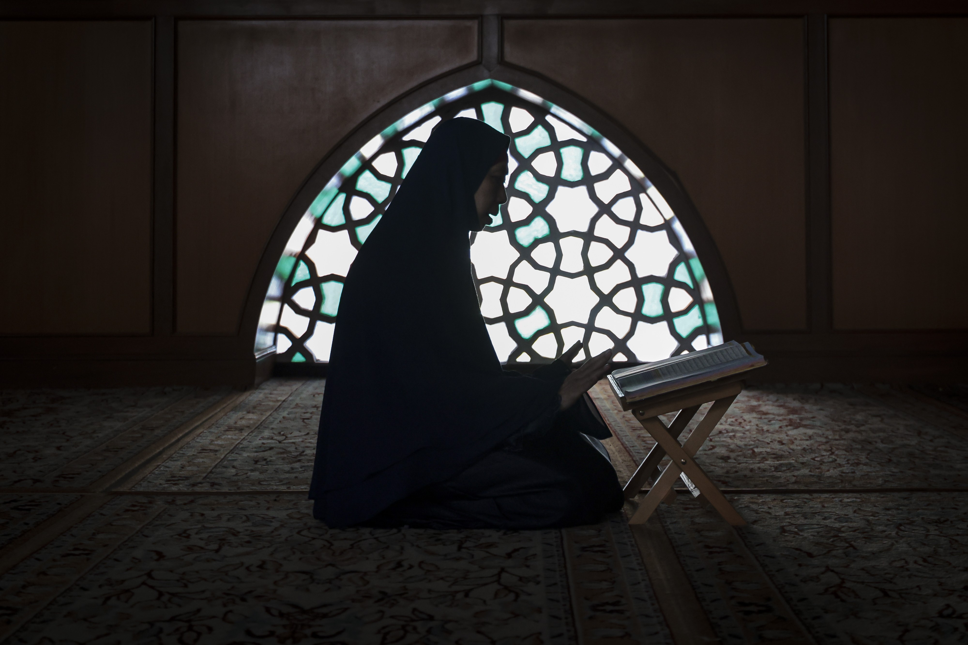 A woman sits on the floor inside a dark room and a mosque, reading a Quran.