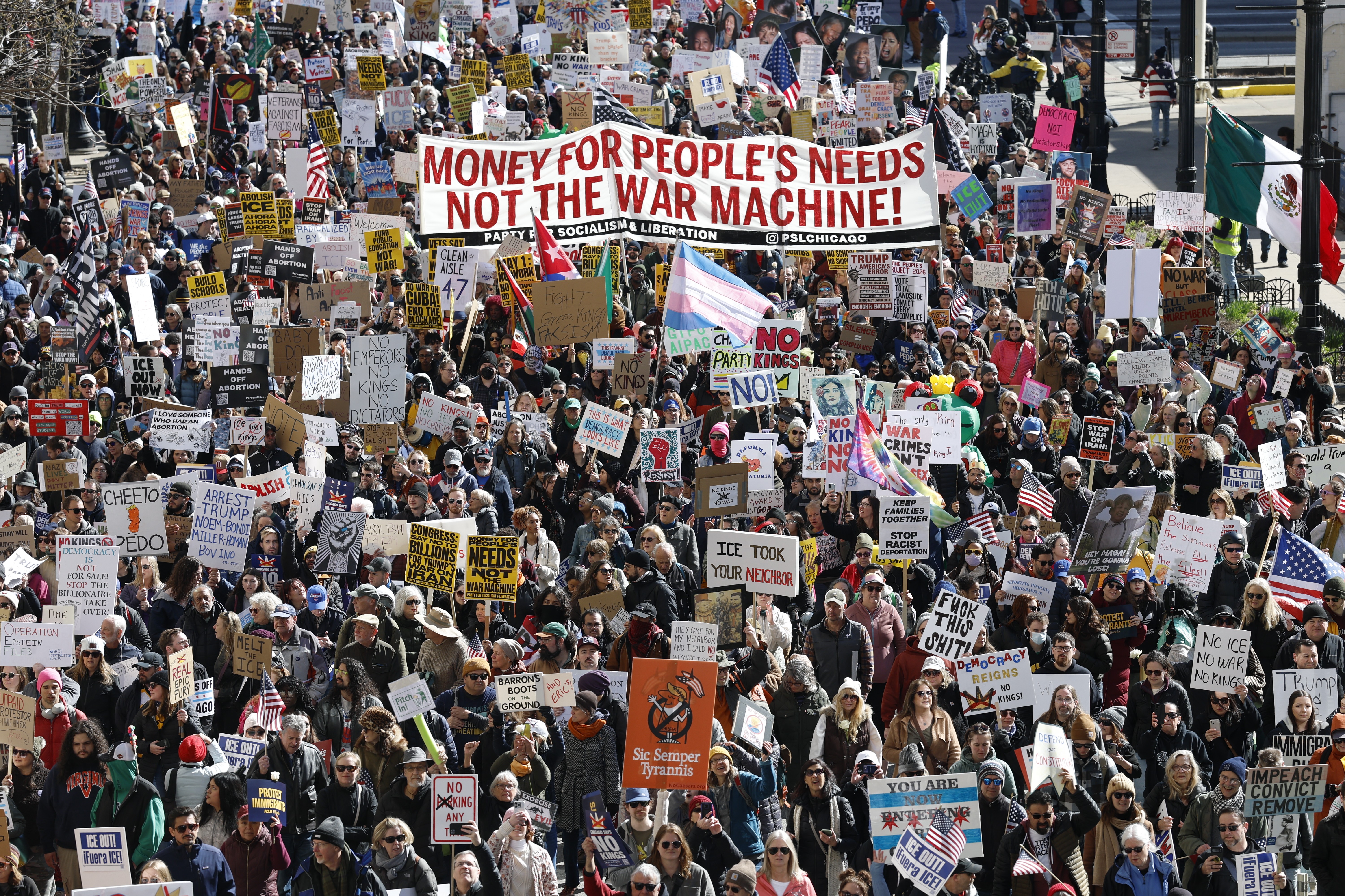 A large crowd of protesters marches down a city street carry many signs and a banner that reads