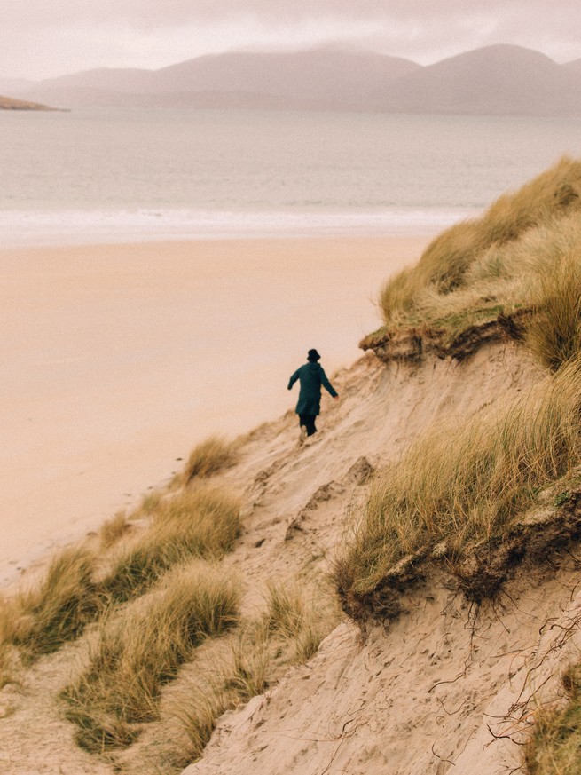Photo of a person in a coat and hat scrambling along steep, grassy dunes next to a broad, sandy beach