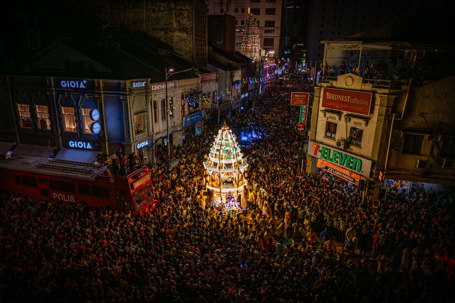A large crowd gathers in a street around an illuminated rig carrying a statue.