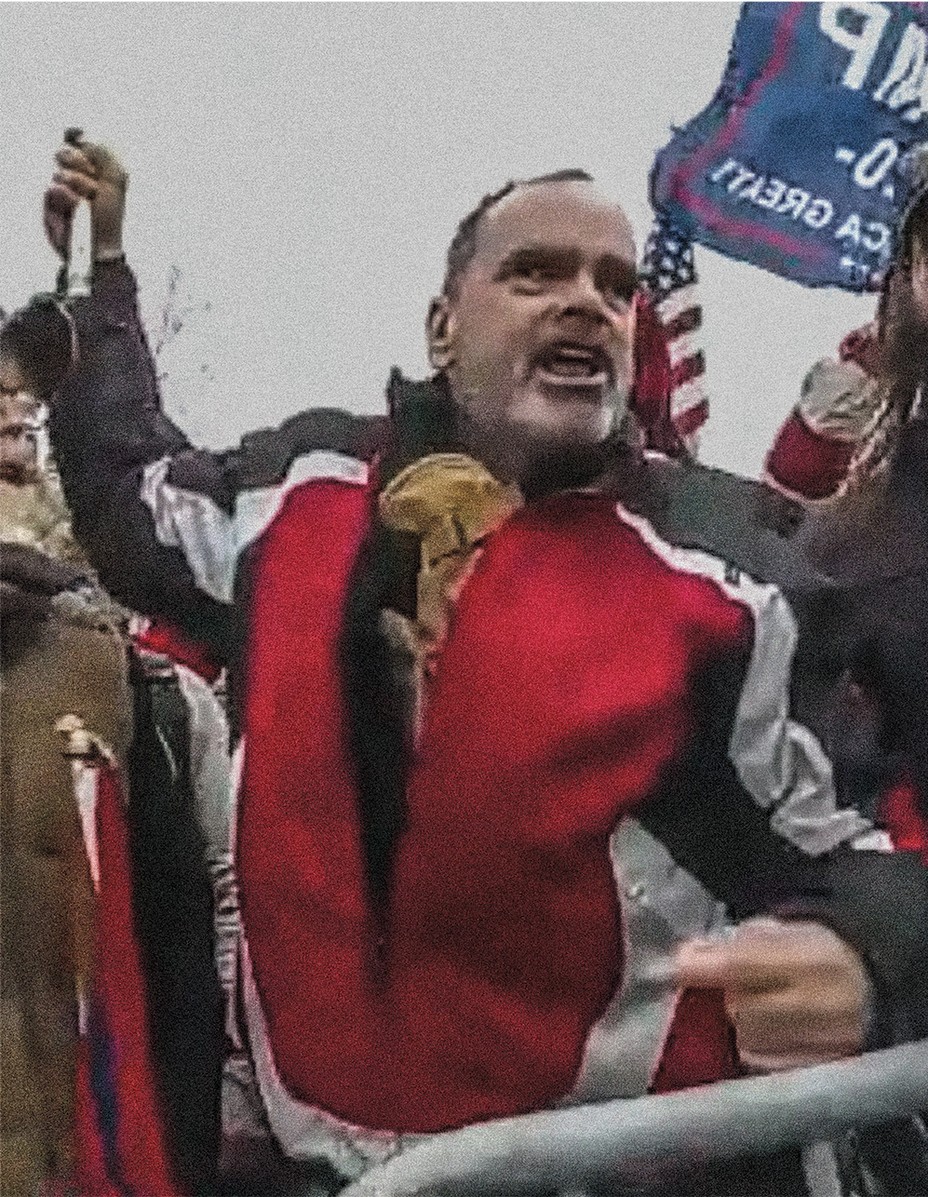 body camera photo of man in red, black, gray jacket raising pole over his head and yelling just on other side of metal barrier