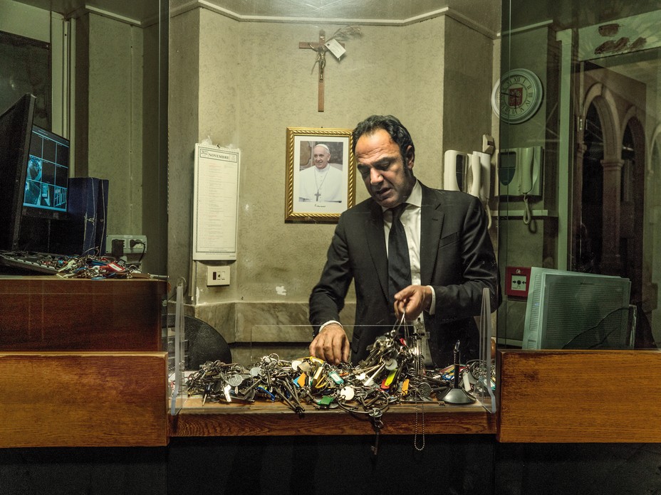 color photo of man in suit sorting enormous keyrings with hundreds of keys in room with photo of Pope Francis and crucifix on wall behind
