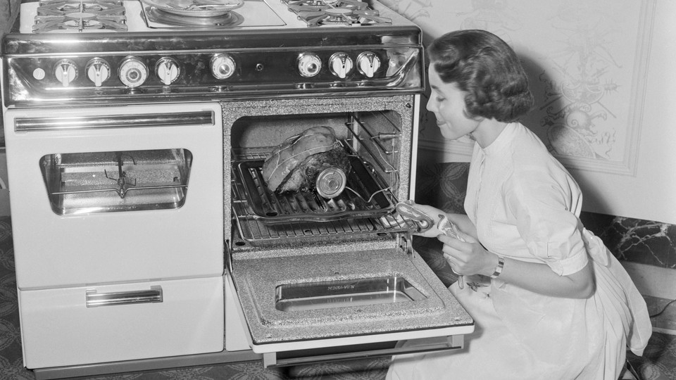 A black-and-white photo of a woman cooking a piece of meat in a gas oven