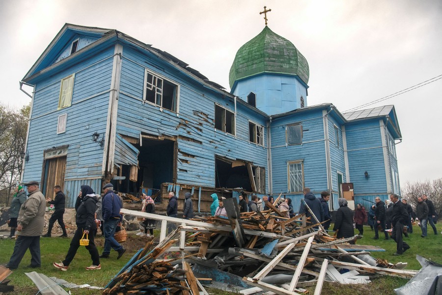 People walk about in the yard of a bomb-damaged wooden church.