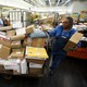 A U.S. Postal Service employee stands next to a cart full of packages.