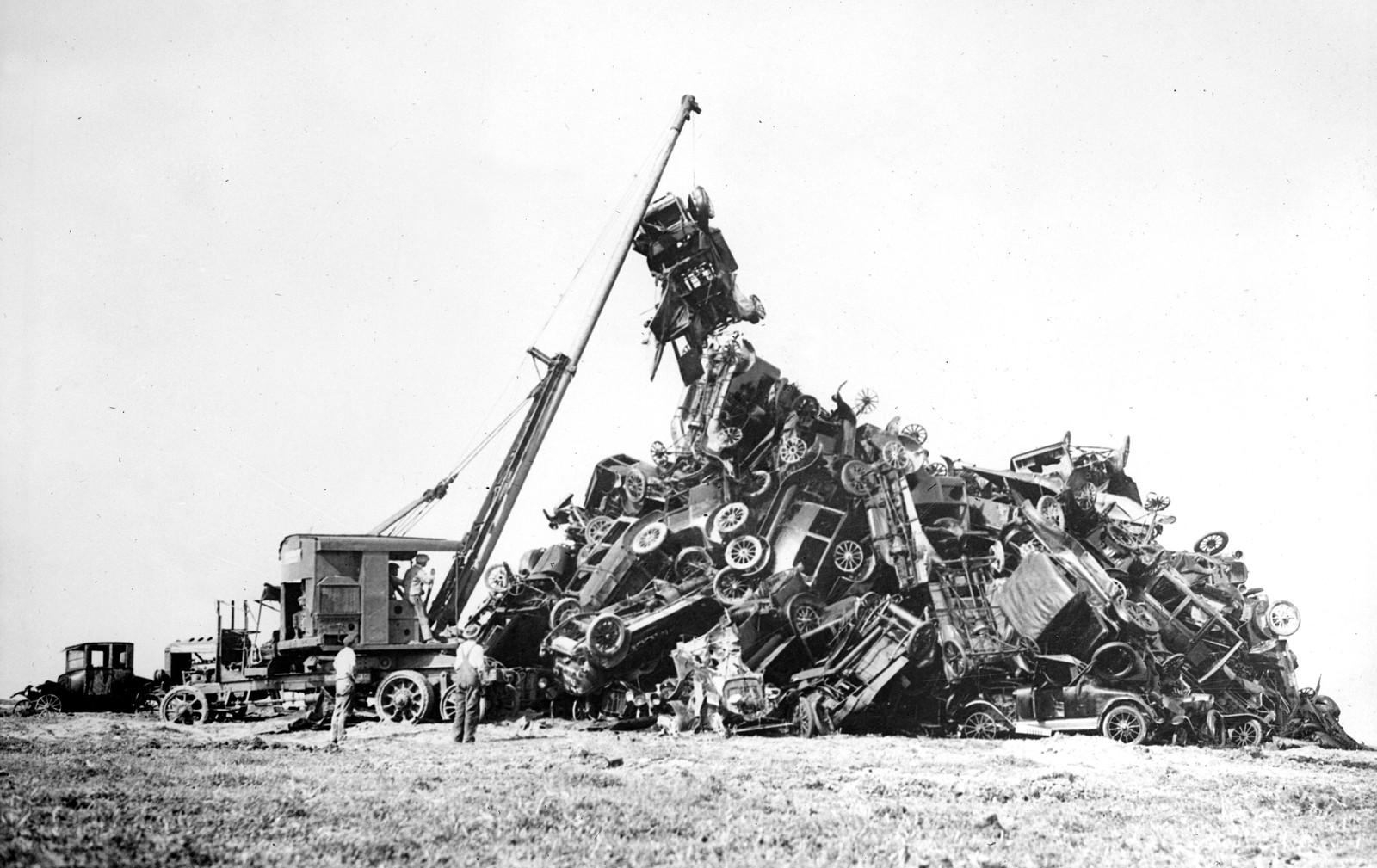 A crane is used to place a wrecked car on top of a large pile of many other wrecked cars, all manufactured around the 1920s.