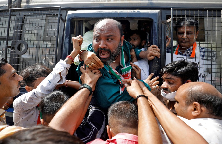An angry man is pushed into a police vehicle by many others below him.