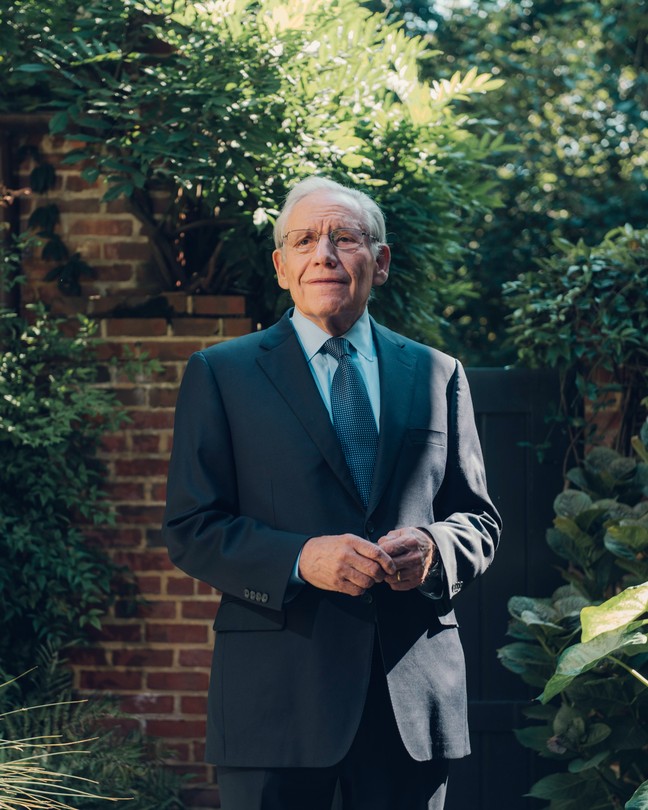 An older man in a blue suit and tie standing in front of a brick wall and lush greenery
