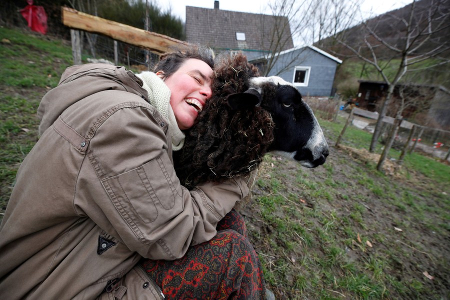 A woman sits on the ground, smiling while hugging a sheep by the neck.