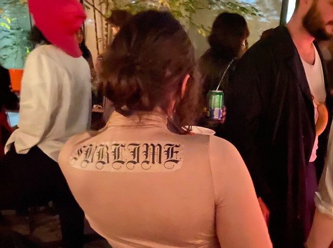 People at a party, a woman's back with a skin-tone bodysuit and a Sublime back tattoo iron-on.