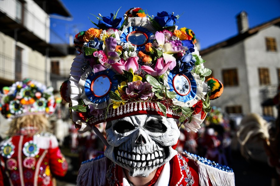 A person wears a skull mask and a large hat covered in flowers and mirrors.