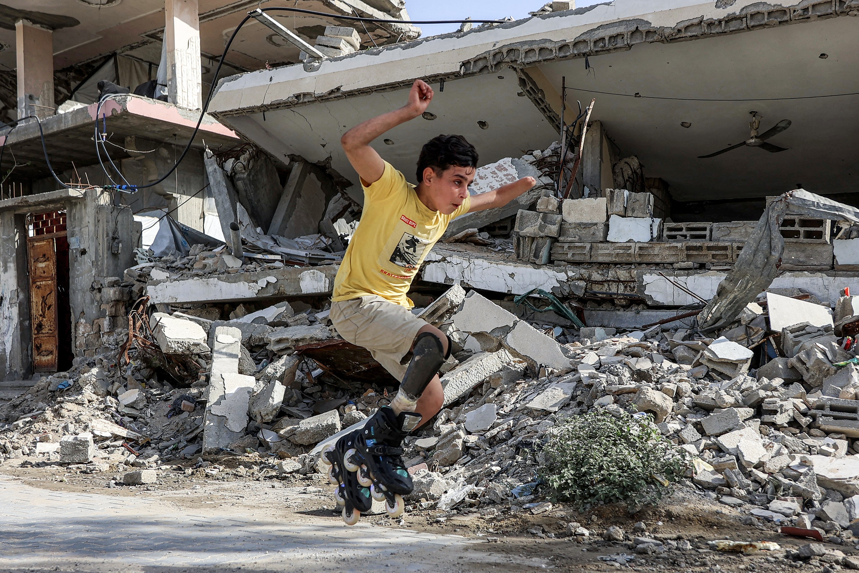 A boy jumps while rollerblading in front of destroyed buildings. The boy is wearing a prosthetic leg attachment as well.