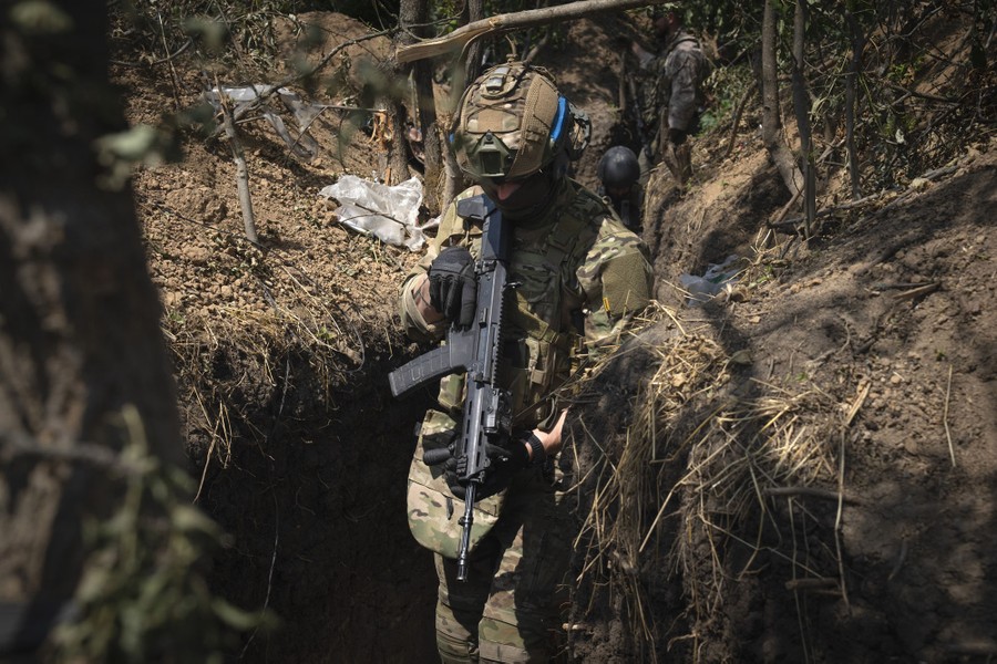 Several soldiers walk in a narrow trench dug in a wooded area.