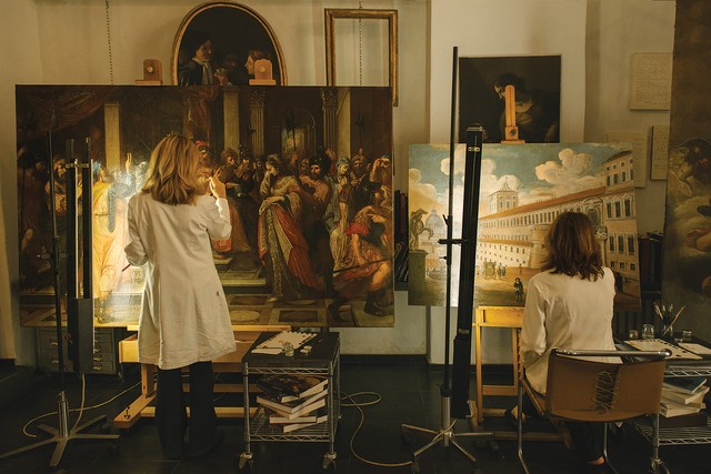 photo of two women from back, one sitting and one standing, while working to restore on large antique oil paintings on easels