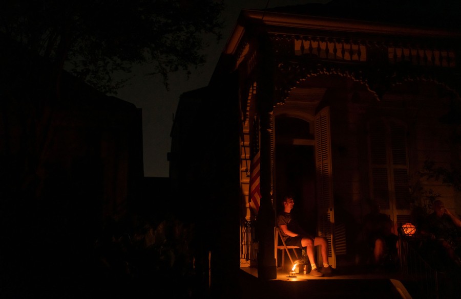 A person sits on their porch at night, lit only by the flame of an oil lamp.
