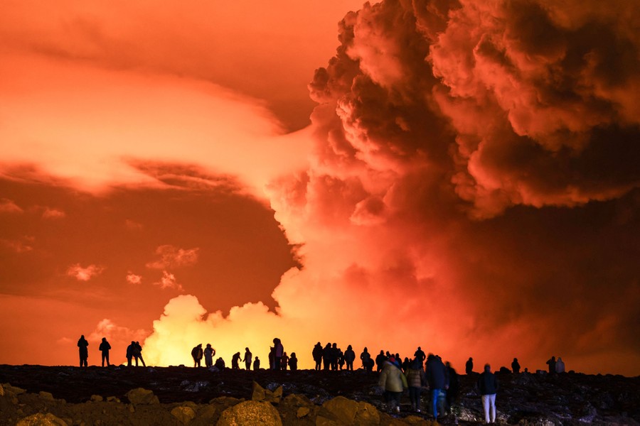 People are seen in silhouette, against a backdrop of steam and gas clouds that are illuminated by glowing lava below.