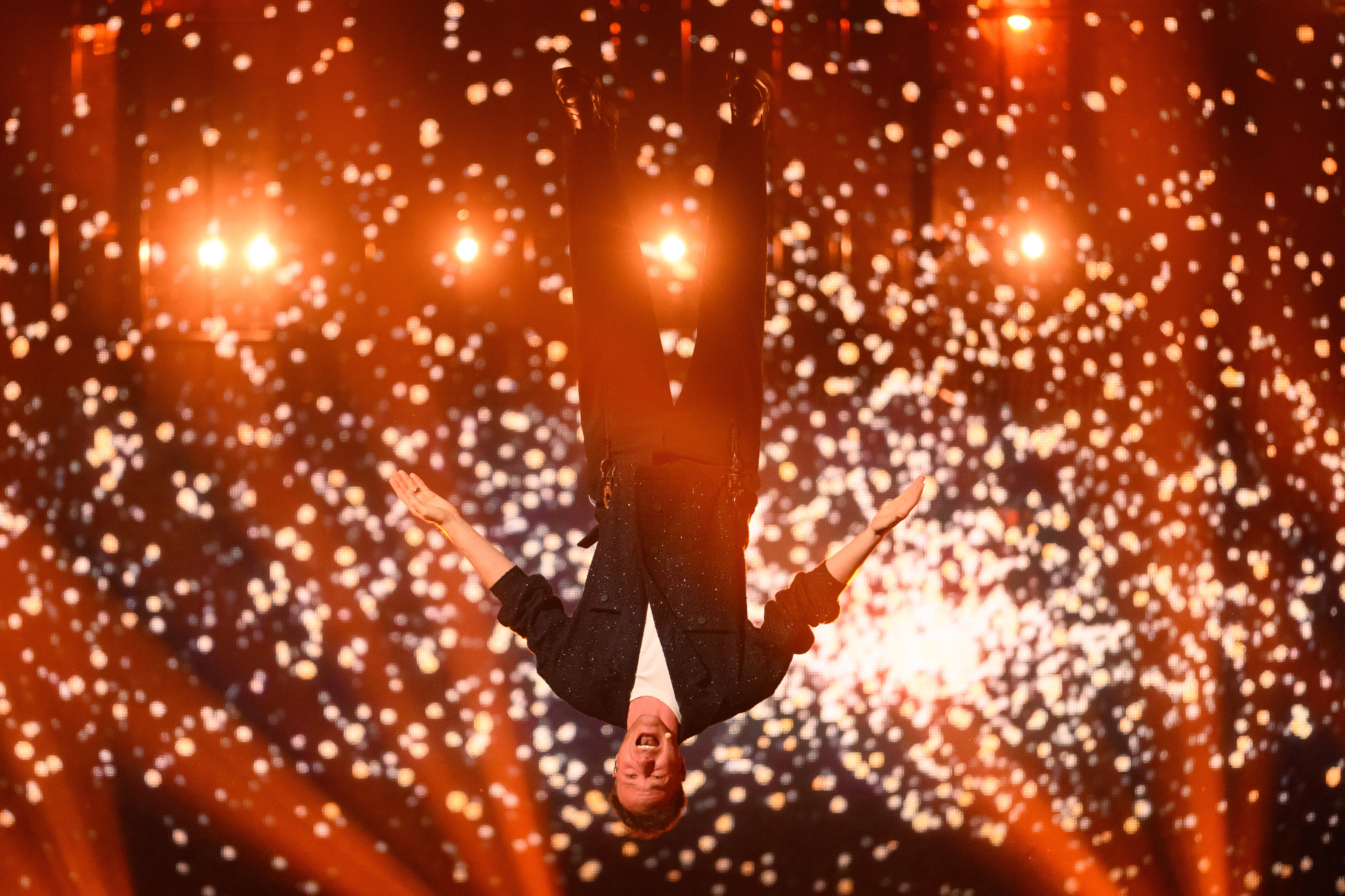 A singer performs while hanging upside down on the stage, sparks cast in the backstage.