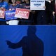 The dark shadow of a man is projected onto a blue barrier. Behind it, attendees at a Trump rally hold signs.
