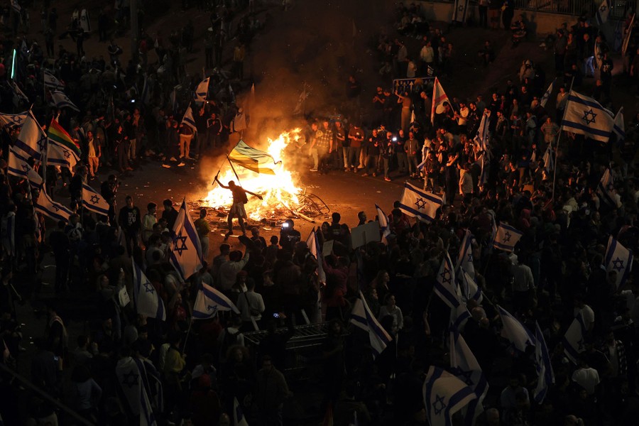 A large crowd of protesters carrying Star of David flags gather around a bonfire.