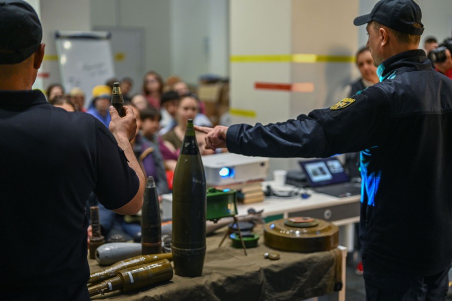 People in a classroom look toward an instructor, who points at a projectile that is sitting on a table full of explosive devices.