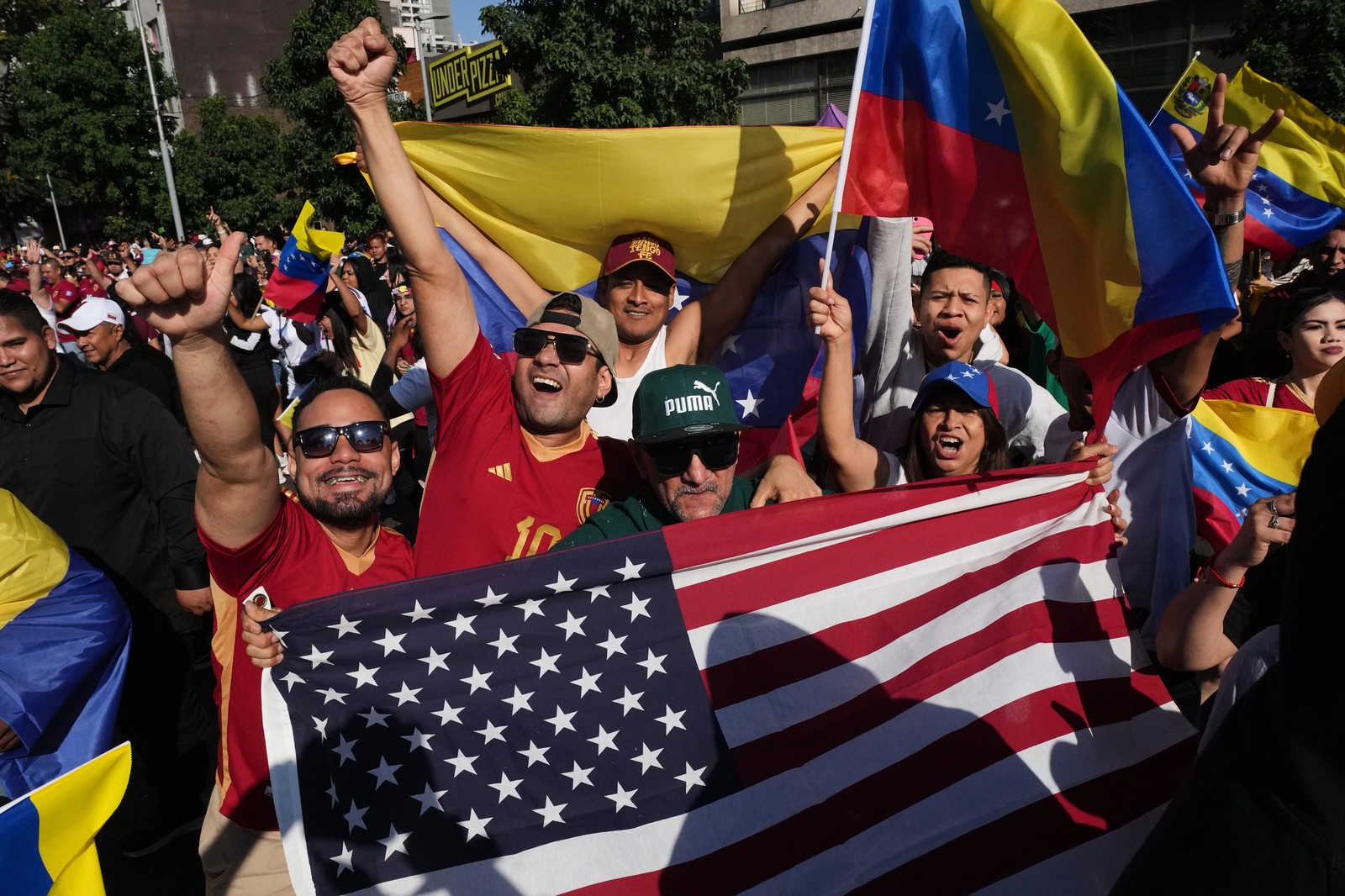 A crowd of people hold Venezuelan flags and an American flag in a public Square.