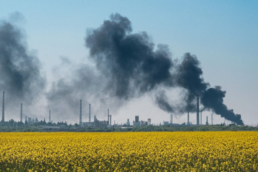 Black smoke rises from a fire above an industrial area seen in the distance behind a flowering field.