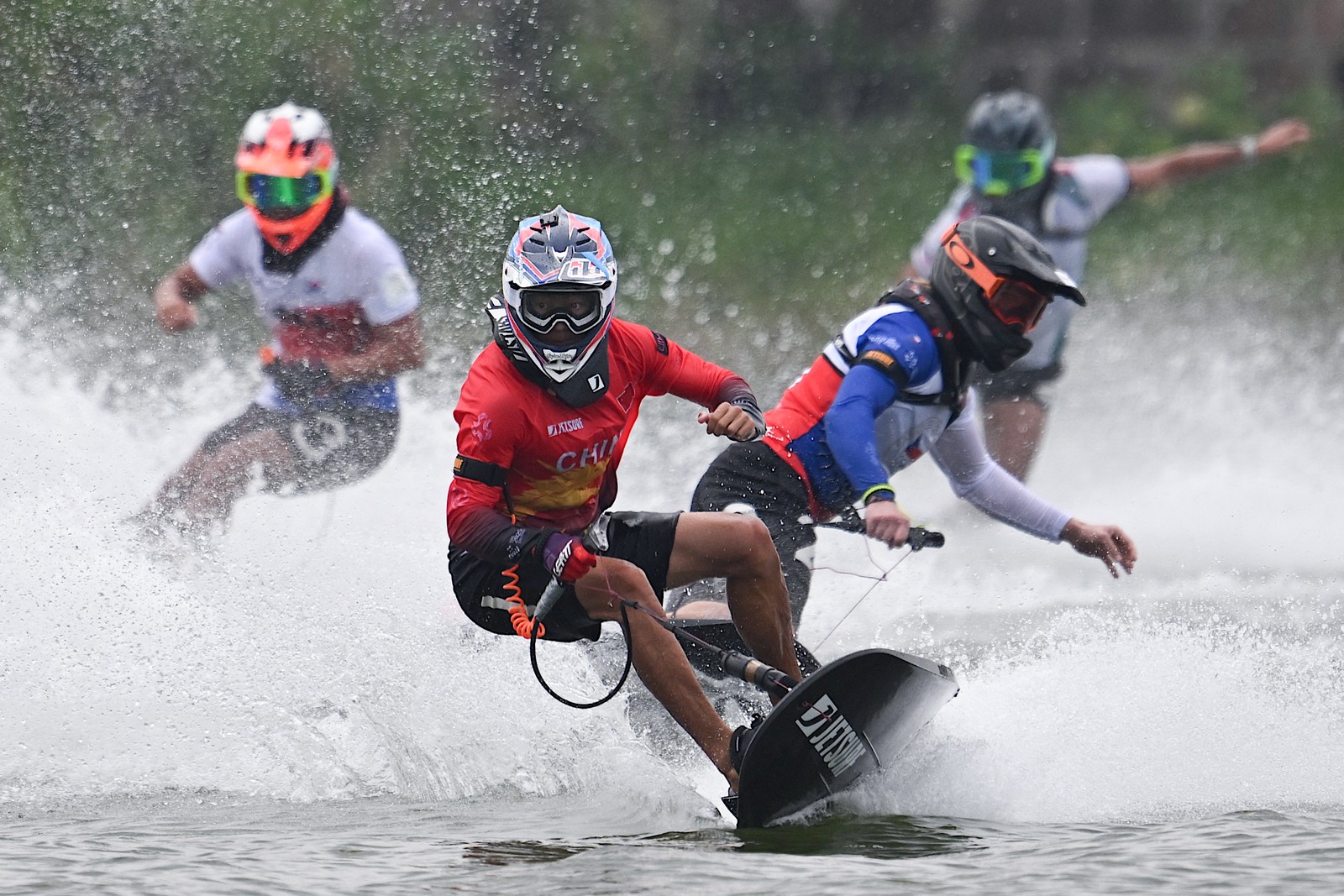 Four people wearing helmets race toward the camera on small motorized boards on a lake.