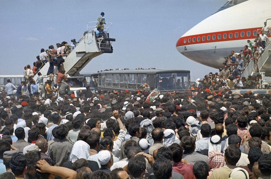 A large crowd gathers beneath and beside an aircraft to welcome people arriving.
