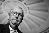 A black-and-white photo of Mitch McConnell seen from below, with a decorative ceiling in the background