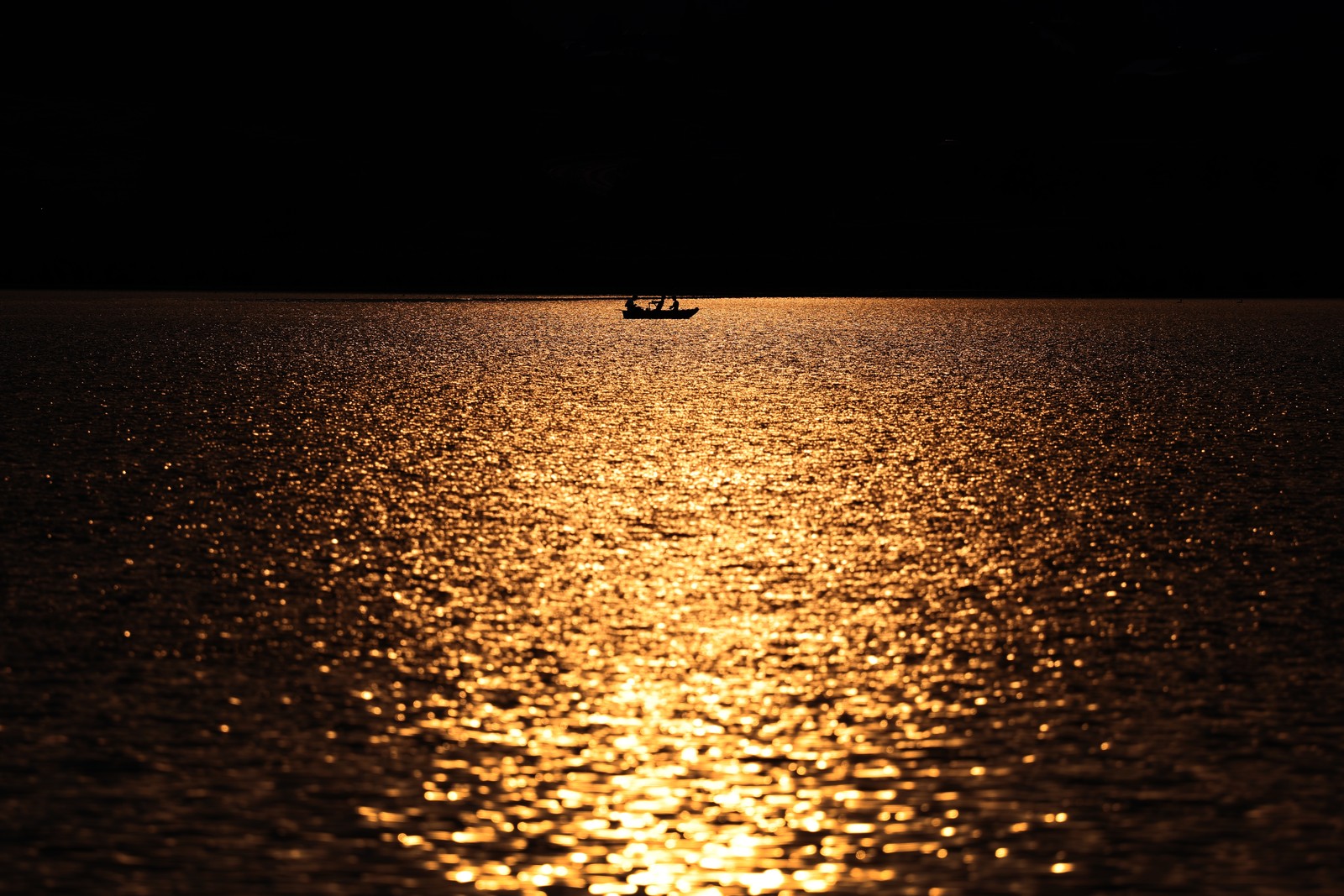 Anglers row a boat across a lake that glistens in the sunset.