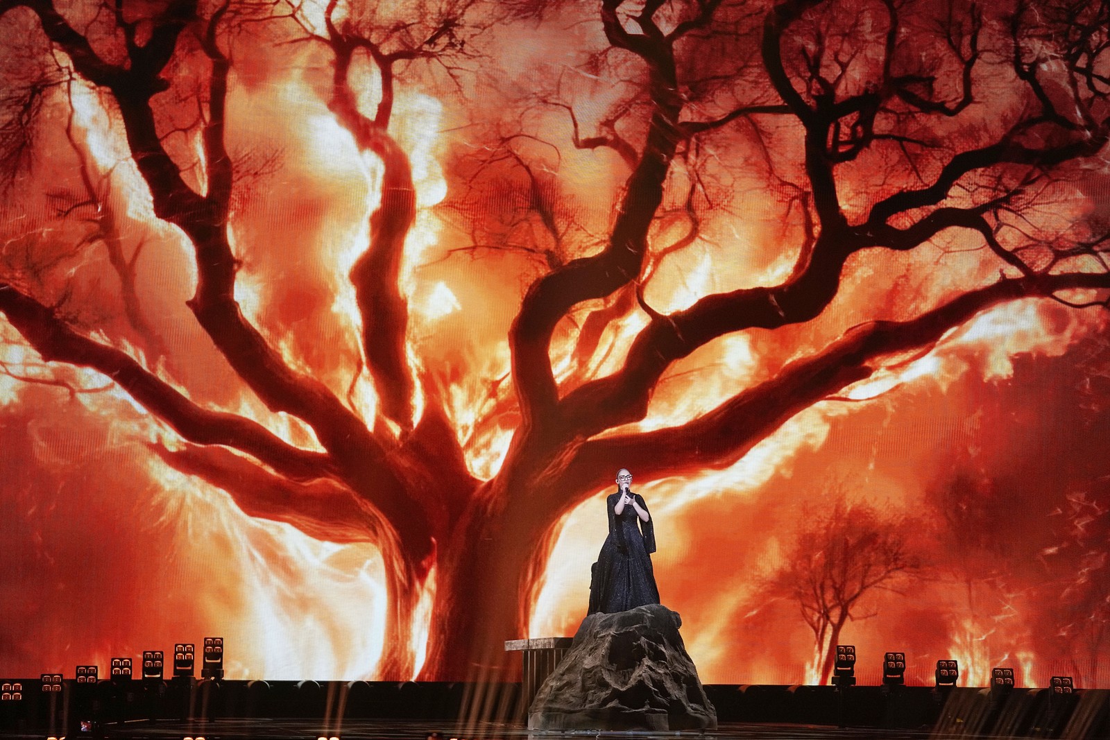 A singer performs on a broad stage in front of a huge video image of a burning tree.