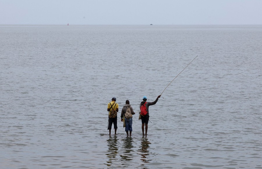 Three men in ankle-deep water attempt to catch fish.