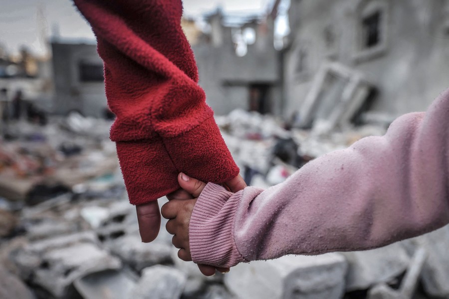 A close view of two people holding hands, with the rubble of a building in the background.
