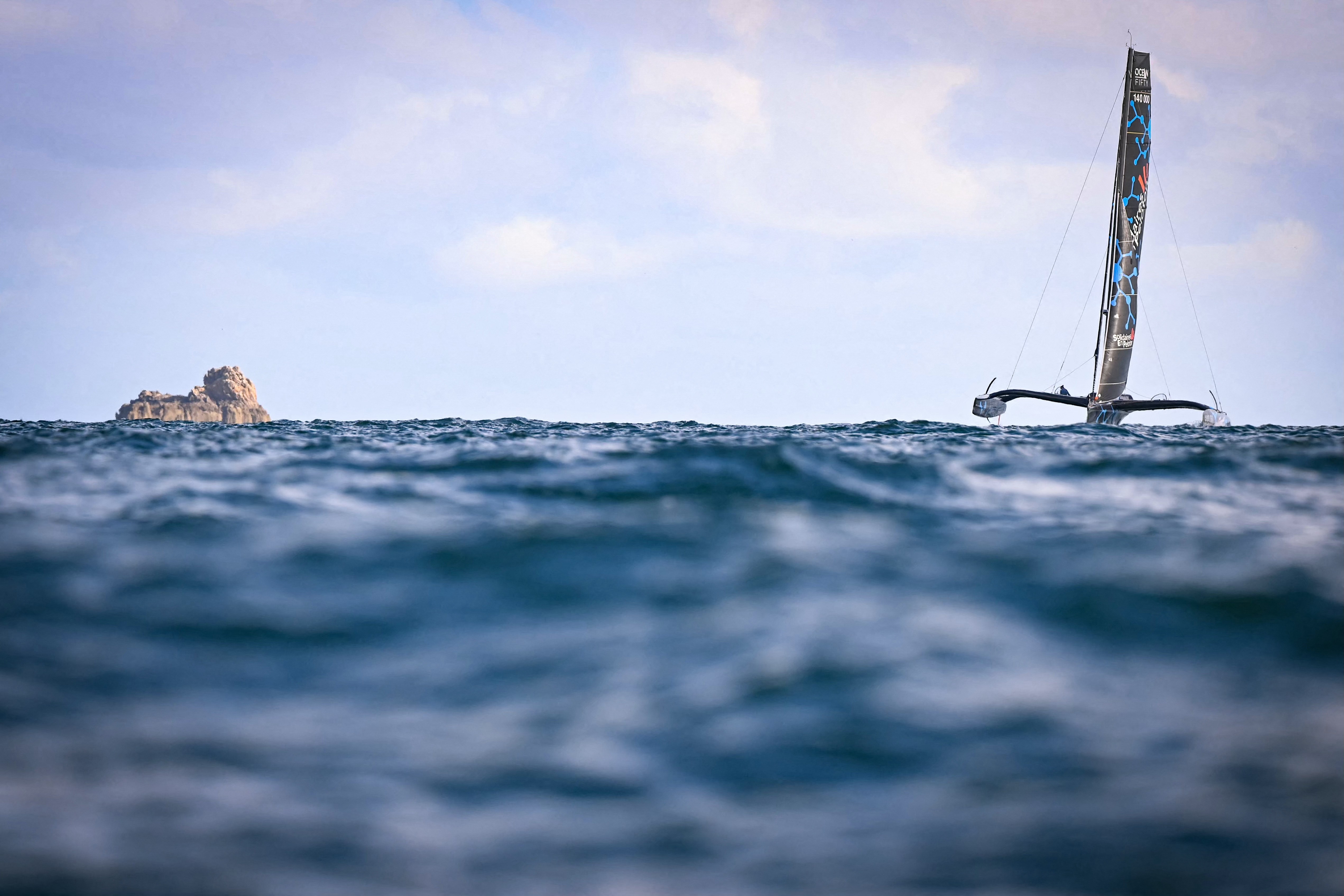 A trimaran, at sail, with a distant rock formation in the background