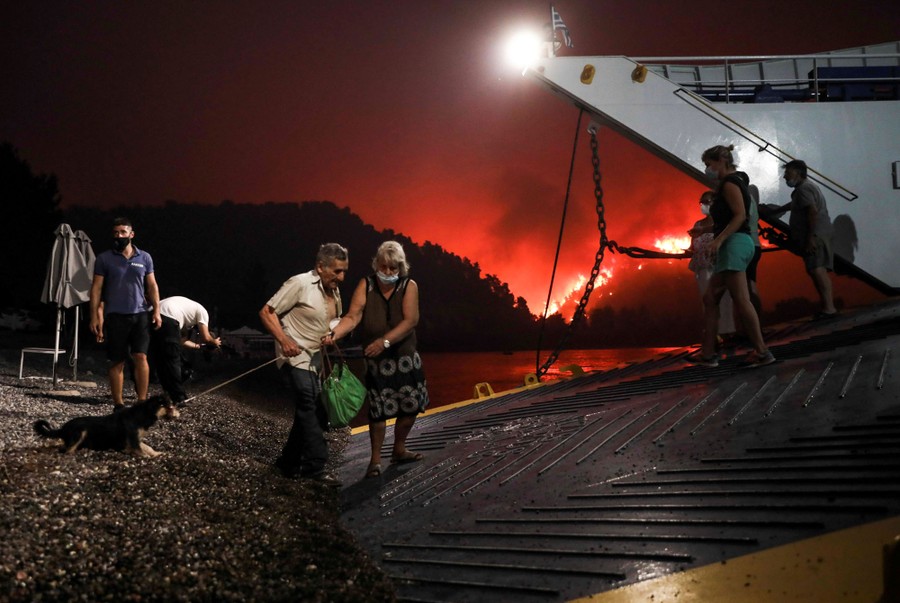 People board a ferry during evacuation as a wildfire burns in the distance.