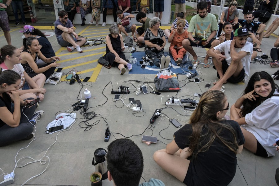 About a dozen people sit on pavement outside a store with their various mobile devices plugged into many outlets.