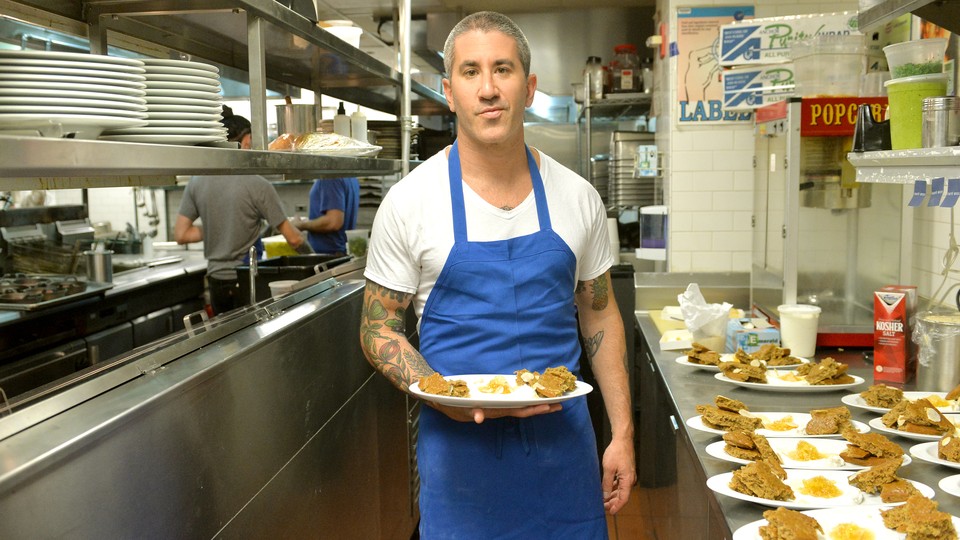 Michael Solomonov holds a plate of food next to a row of plates of food on a restaurant kitchen counter.