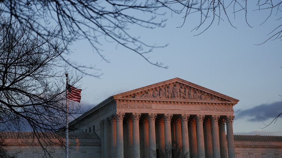 The Supreme Court (and American flag next to it) are slightly pink in a sunset, and framed by leaf-less trees.