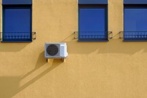 An AC unit mounted on a yellow wall that has three blue windows