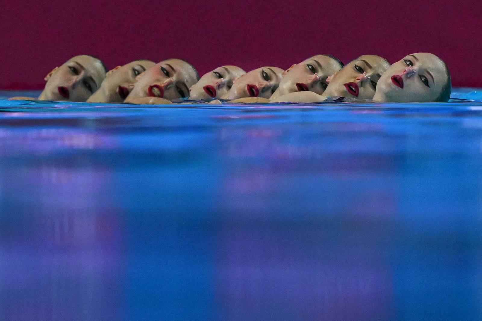 Eight artistic swimmers in a team are seen at the water's surface, all leaning back during a performance.