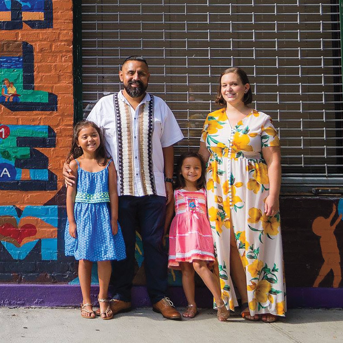 photo of man in guaybera and pants, woman in floral dress, and two girls also in sundresses smiling on sunny day next to wall