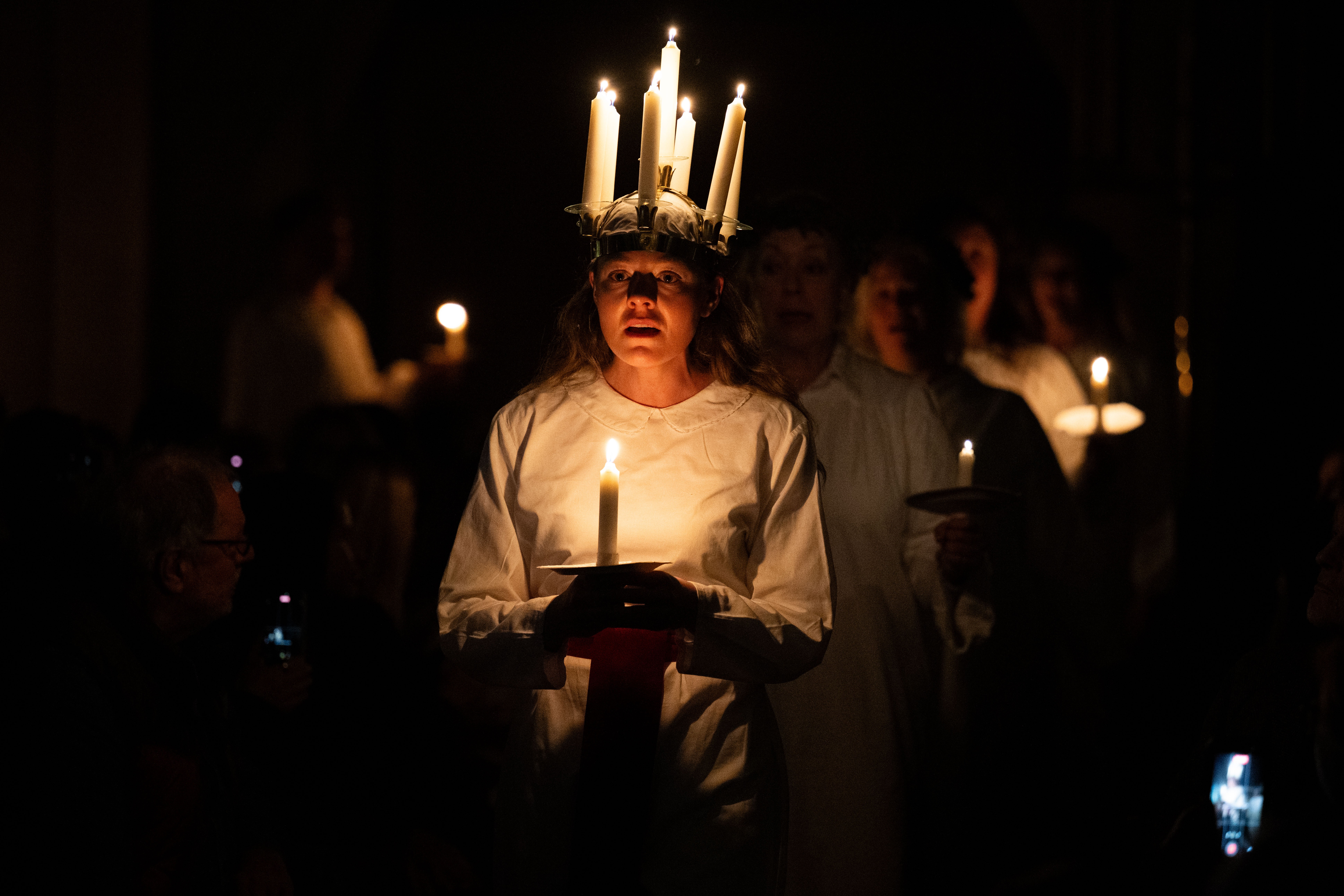 People carry candles during a choir performance, with the person at center wearing a crown of lit candles.