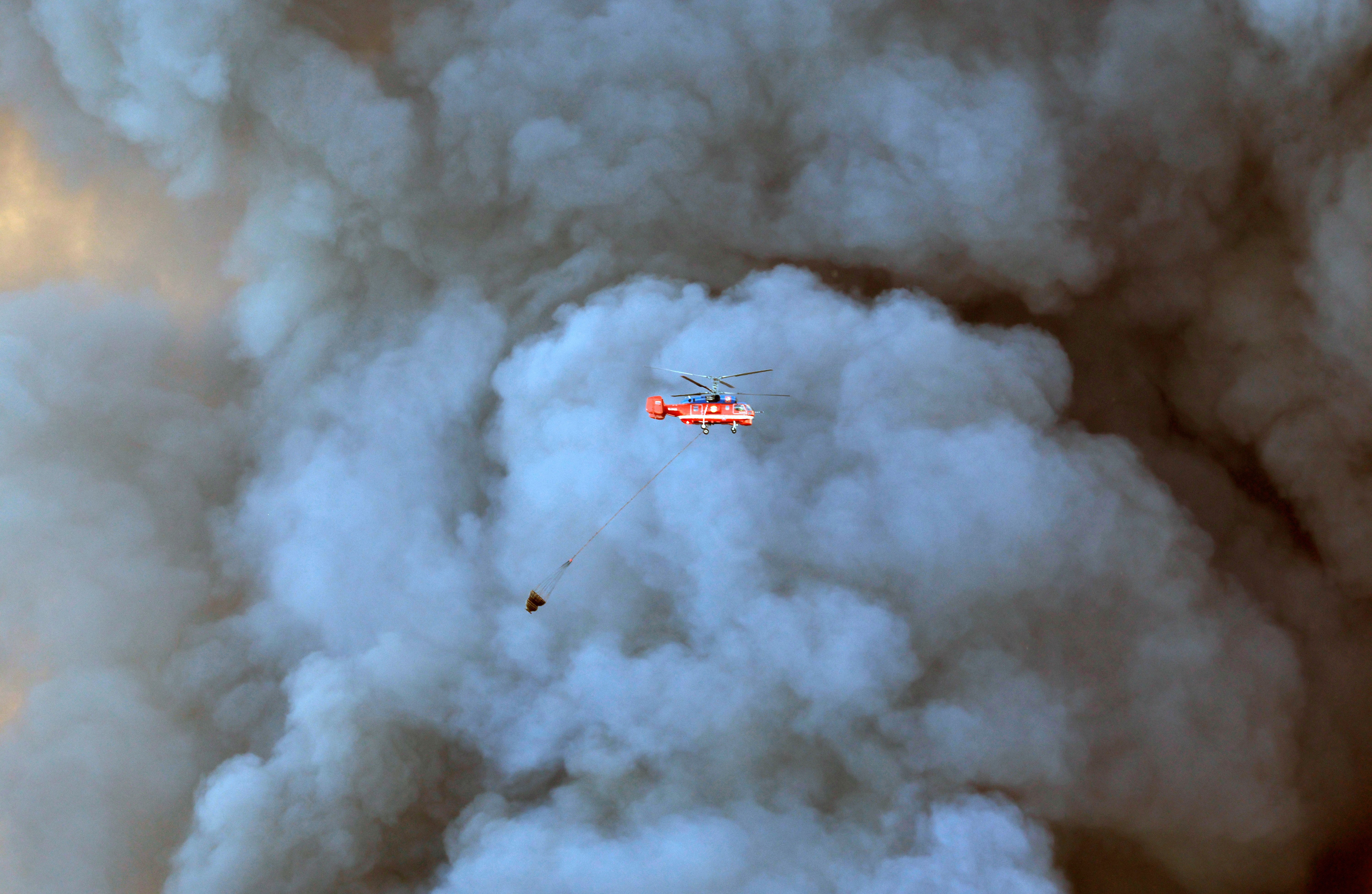 A firefighting helicopter carries a bucket of water past a large cloud of smoke.